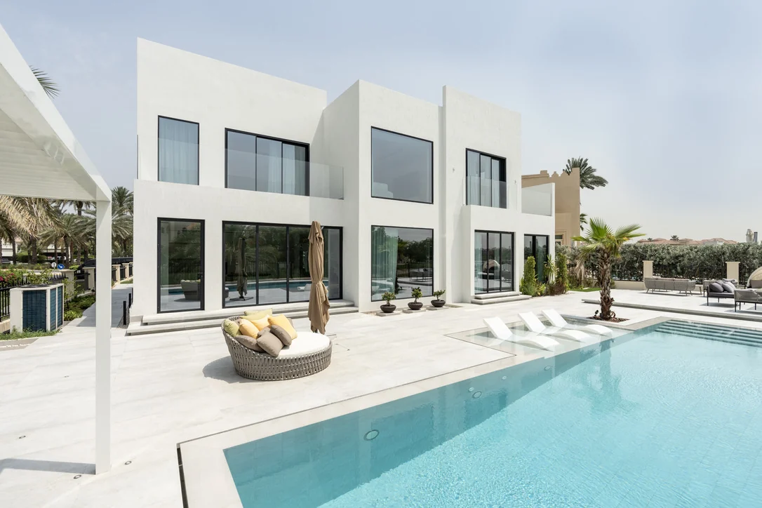 Photo: a white two-storey villa with black-framed glazing, behind a pool and a stone terrace