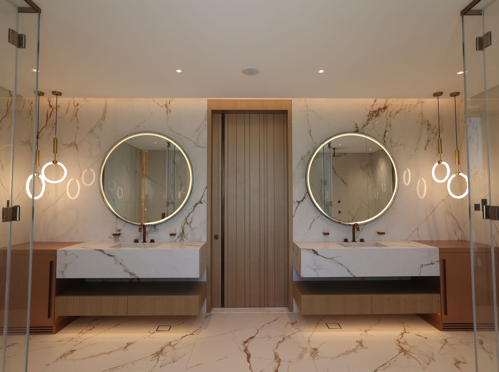 Principal bathroom with book-matched marble walls and floor, twin timber vanities, lit round mirrors and brass pendants, delivered by Fajr Interiors, Dubai