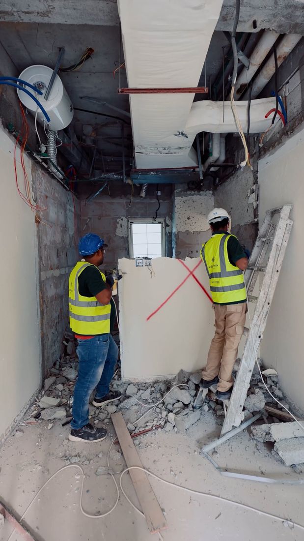 Strip-out at Cluster 49, Villa 13: two Fajr workers in hi-vis opening a wall under the exposed ceiling services