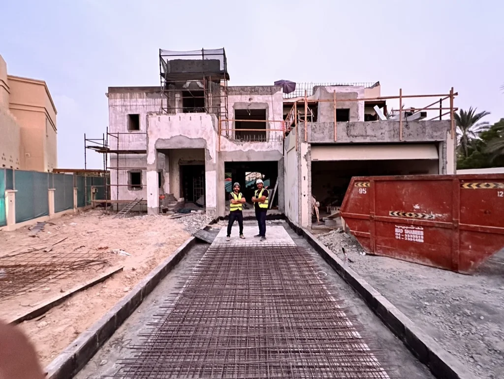 Photo: the front of the villa during the works, stripped to its shell with scaffolding up and rebar laid for the driveway, two site staff in hi-vis on the drive, Cluster 39, Villa 1, Jumeirah Islands