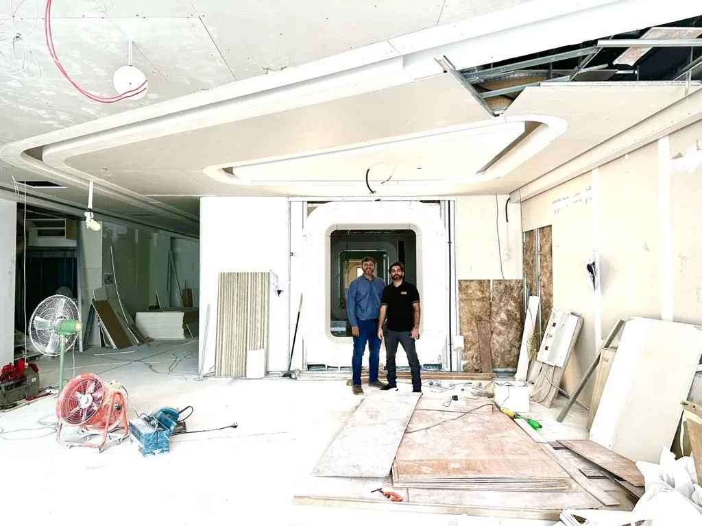 Photograph: Two people standing in a framed portal opening on an office floor under construction, beneath a curved ceiling bulkhead