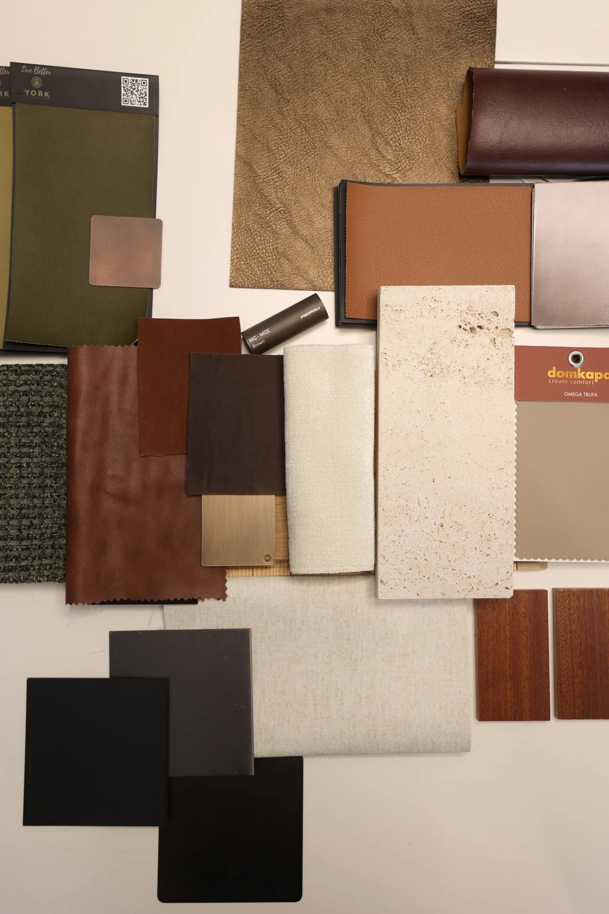 The Opus by Omniyat material board from above: black and bronze metal plates, a brushed brass plate and a bronze chair-leg finish among leathers, travertine and veneer